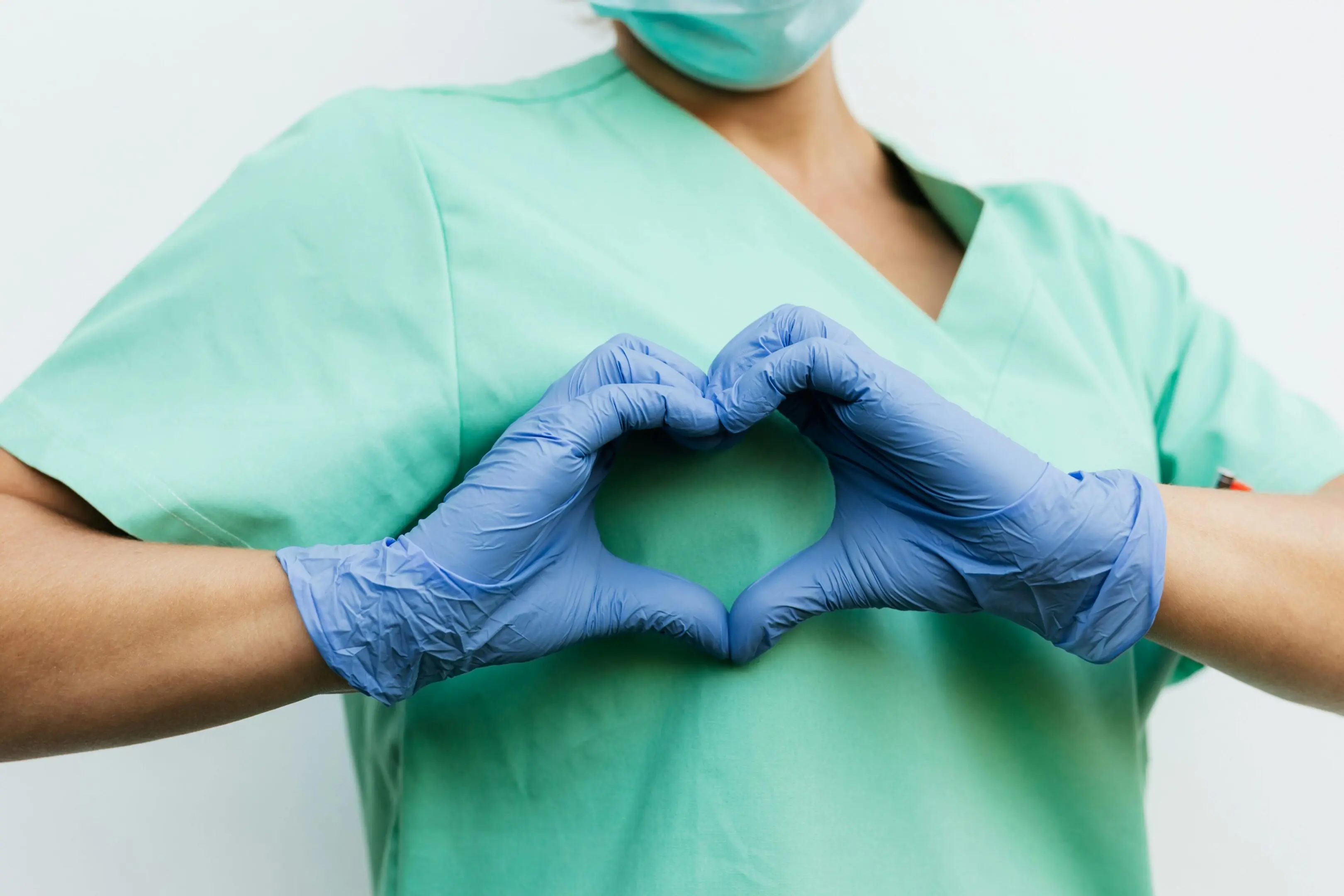 Healthcare worker in scrubs and gloves making a heart shape with hands.